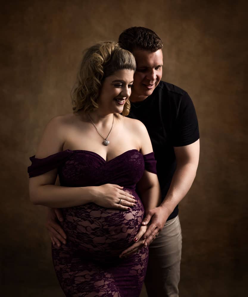 a moody colour maternity photograph of a pregnant lady in a wine coloured lace gown with her partner embracing her